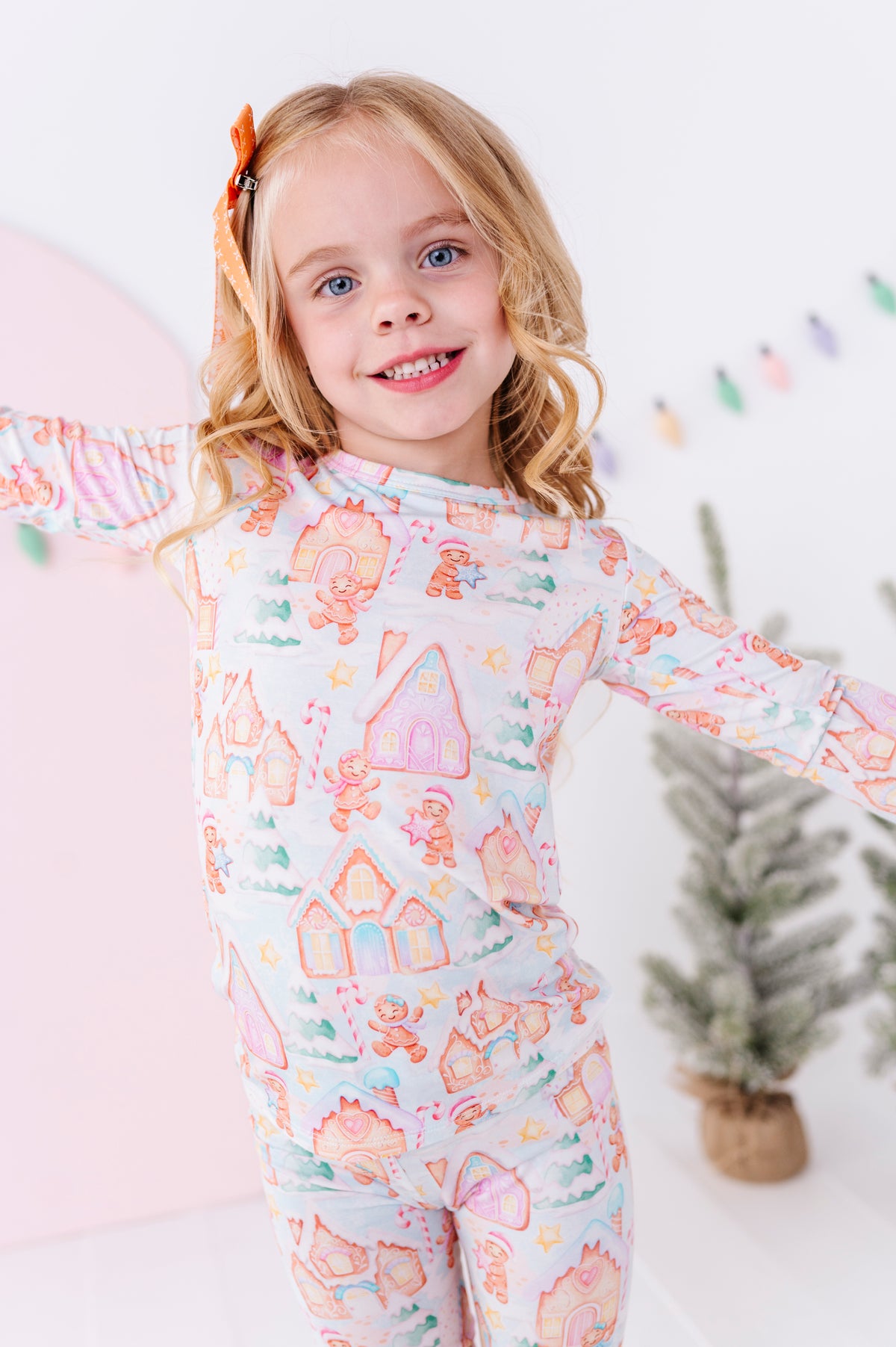 A young girl with blonde hair stands in a bright white room wearing a long-sleeve two-piece bamboo pajama set with a pastel gingerbread village print.  