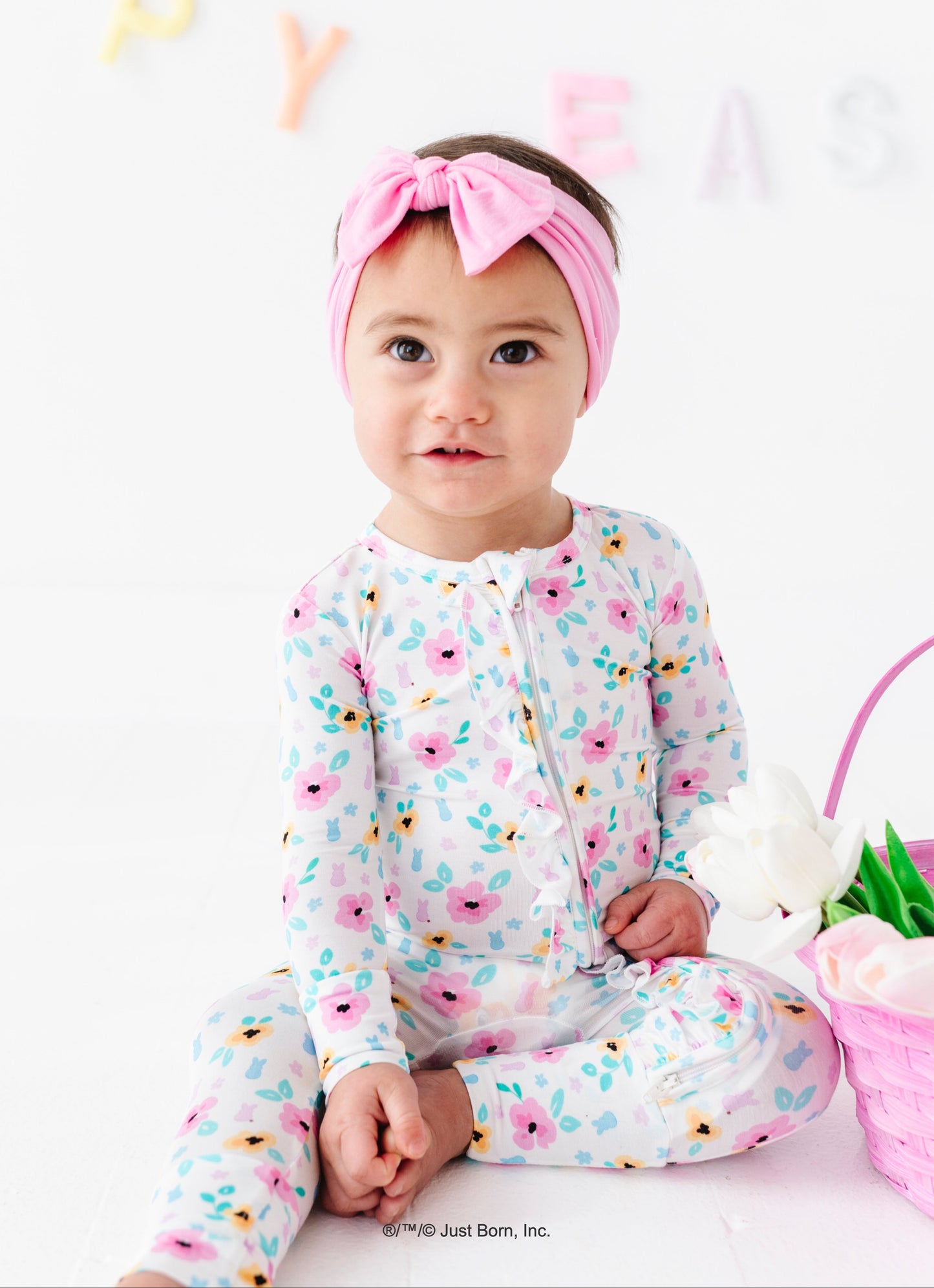 Baby wearing a floral PEEPS® bamboo romper with a pink headband, sitting on a white surface with a colorful background.