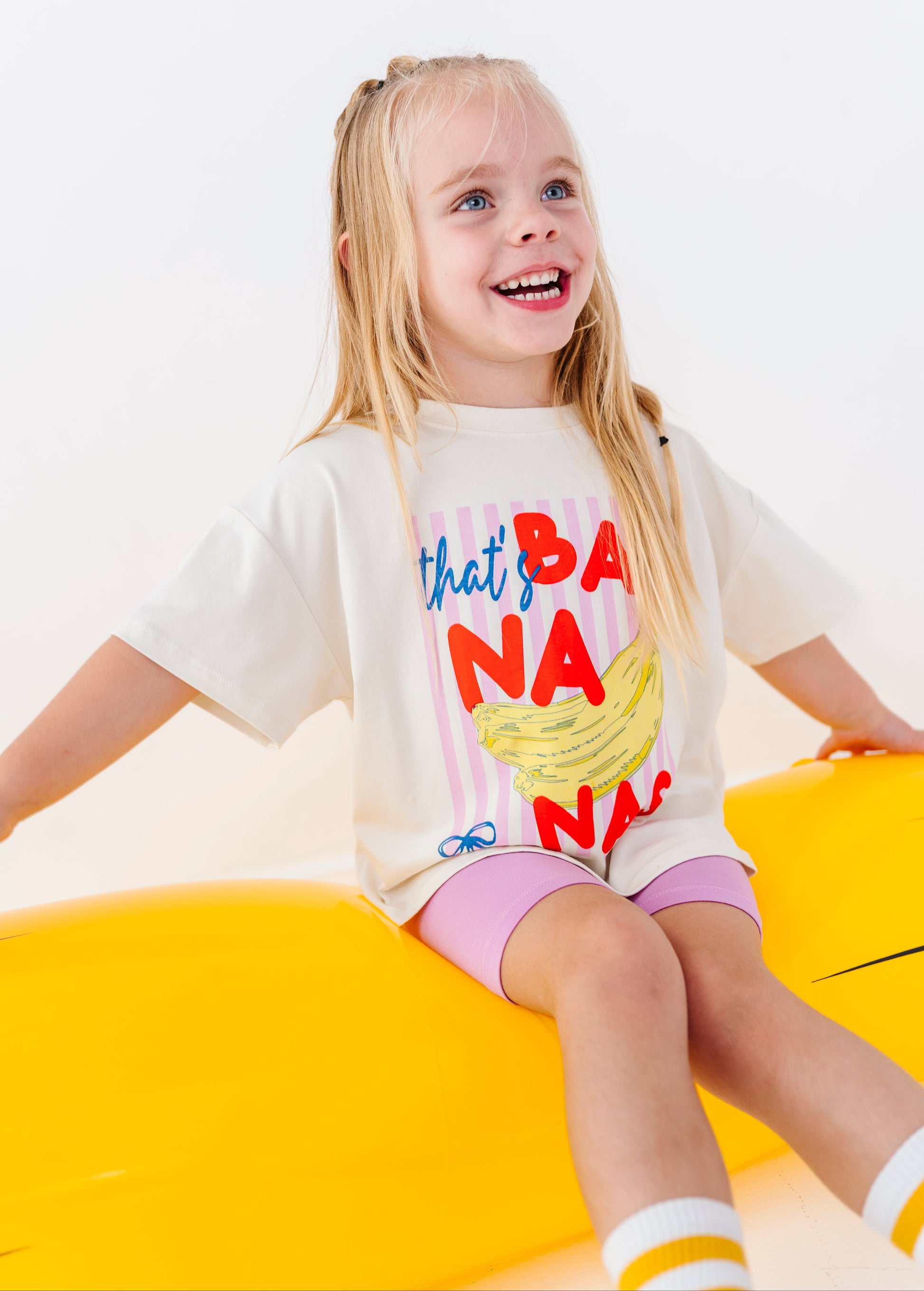 A young girl with blonde hair sitting on a bright yellow float wearing a white graphic t-shirt that reads 'that's BANANA' in red, blue, and yellow text, paired with pink shorts.