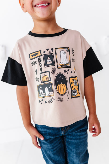 A child wearing a short-sleeve t-shirt with a beige body, black sleeves, and a black crew neck. The front of the shirt features a Halloween-themed graphic print with ghosts, pumpkins, bats, and stars.