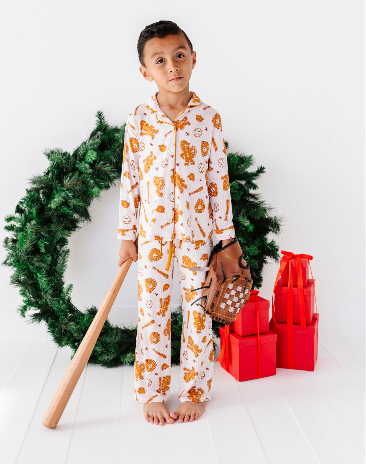 A young boy stands barefoot in a holiday-themed studio setting, wearing a white long-sleeve pajama set with an orange gingerbread cookie print. He holds a wooden baseball bat and a small baseball glove, positioned in front of a green Christmas wreath and red gift boxes on a white wooden floor.