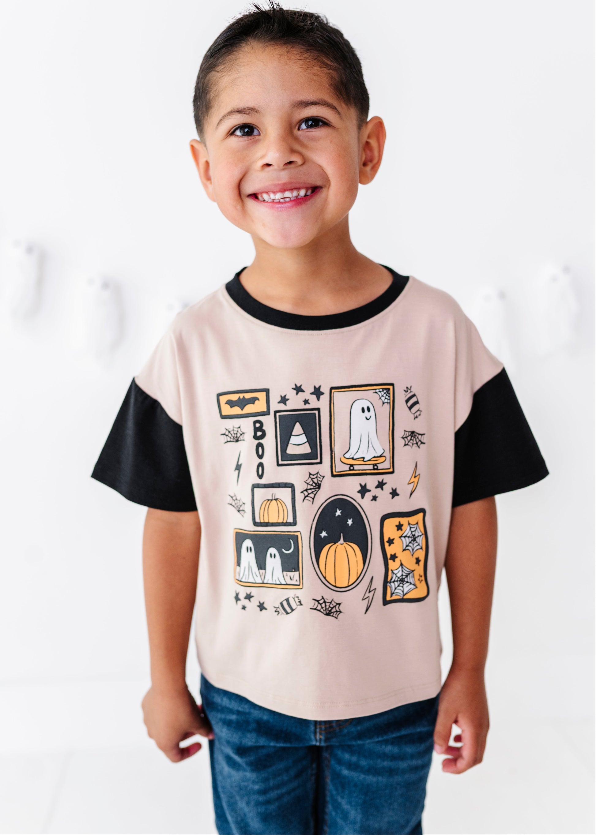 A smiling young boy wearing a light beige t-shirt with black sleeves and a colorful Halloween graphic print, paired with blue jeans, standing against a white wall.