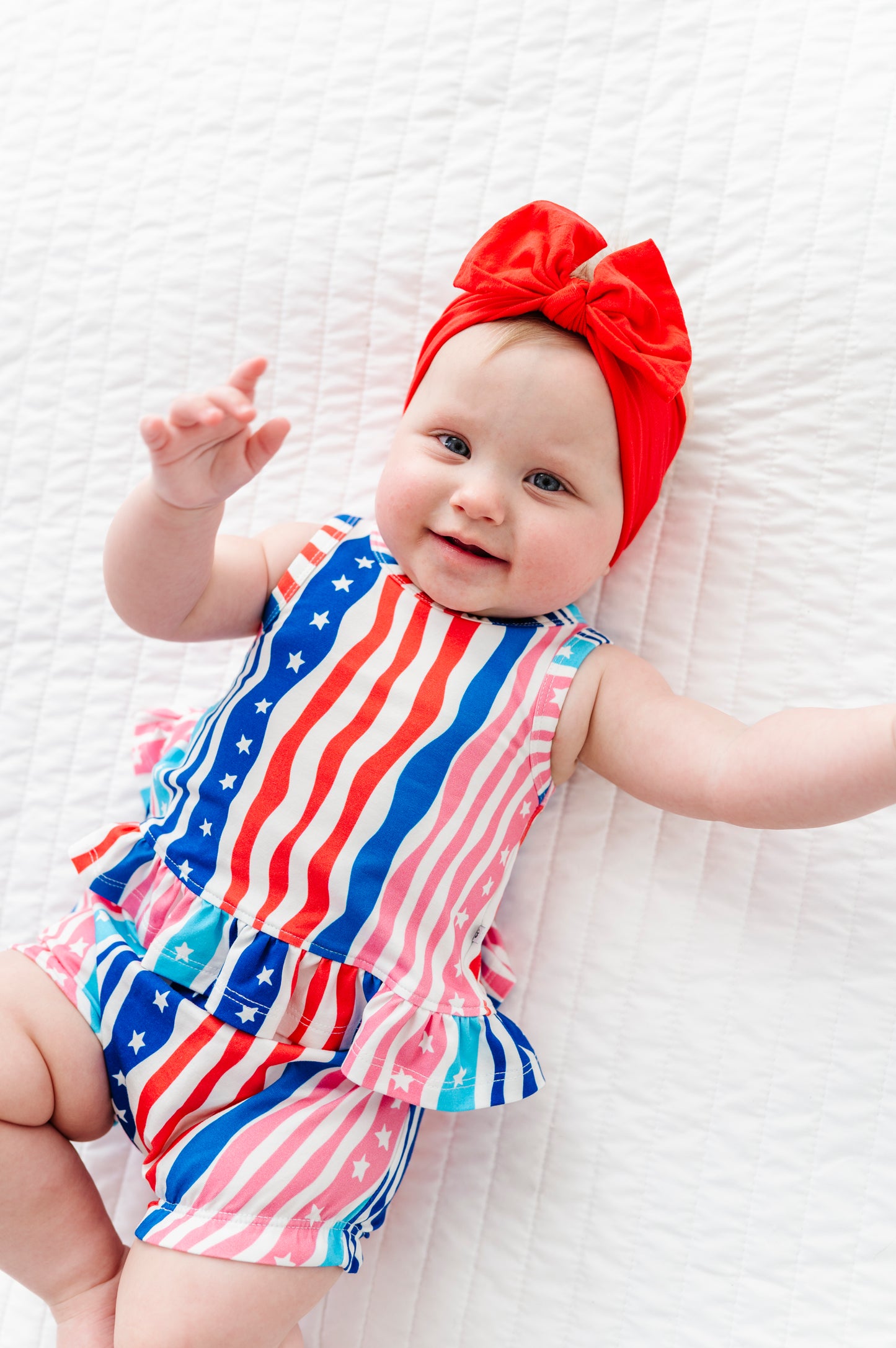Baby wearing a colorful American flag outfit and red headband on a white textured surface