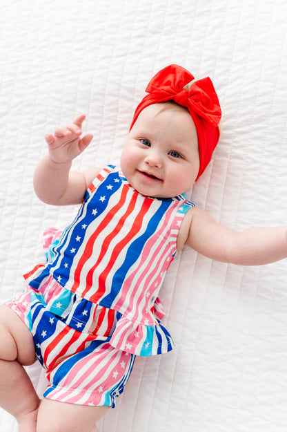 Baby wearing a colorful American flag outfit and red headband on a white textured surface
