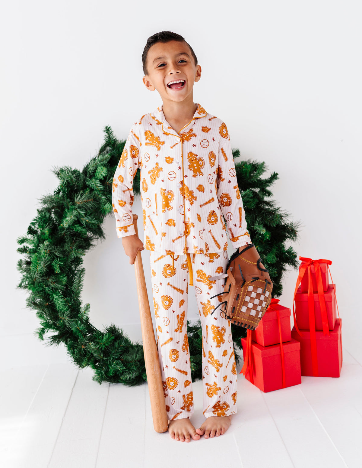 A young boy wearing a white Christmas-themed pajama set with orange and yellow holiday illustrations stands barefoot in a studio setting. He is holding a wooden baseball bat and a brown baseball glove, with a green wreath and red gift boxes behind him.