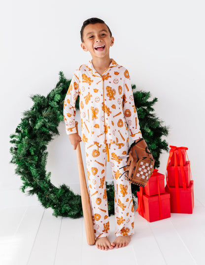 A young boy wearing a white Christmas-themed pajama set with orange and yellow holiday illustrations stands barefoot in a studio setting. He is holding a wooden baseball bat and a brown baseball glove, with a green wreath and red gift boxes behind him.