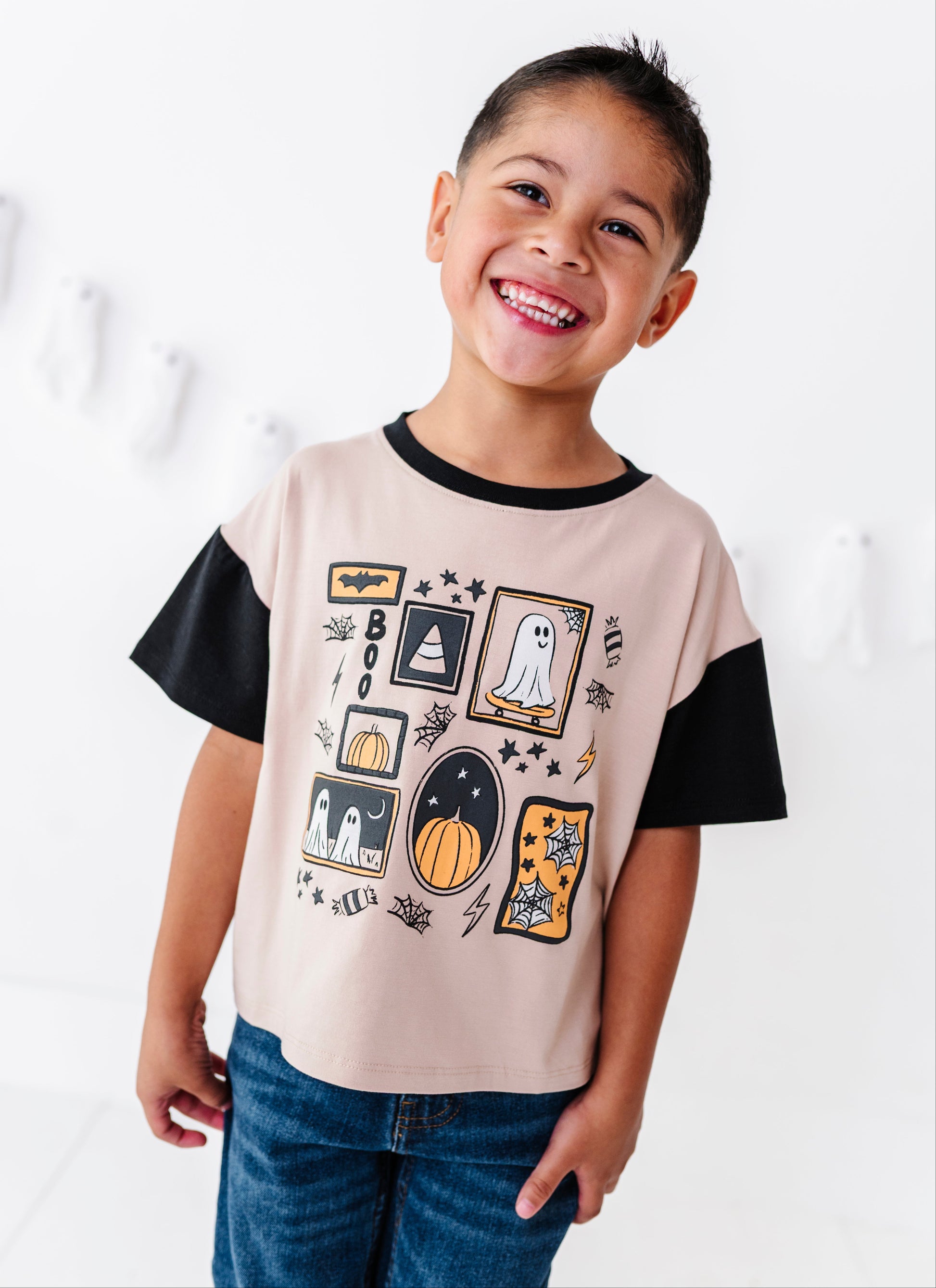 A smiling young boy wearing a beige and black short-sleeve t-shirt with a colorful Halloween graphic print, paired with blue jeans, standing against a white wall.