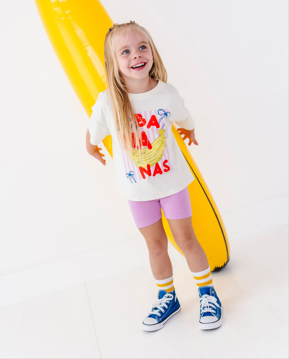 A young girl stands smiling in front of a giant yellow banana-shaped inflatable pool float on a clean white studio background. She is wearing a white graphic T-shirt with red and yellow banana text, pink shorts, yellow and white socks, and blue sneakers.