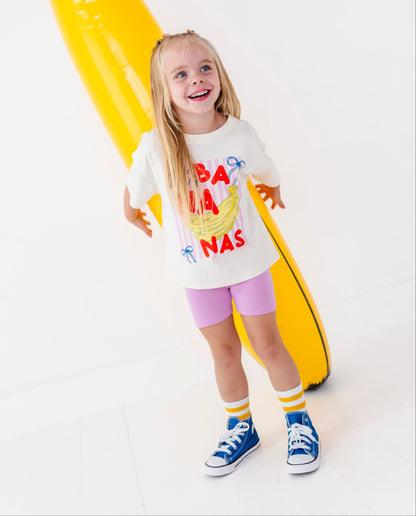 A young girl stands smiling in front of a giant yellow banana-shaped inflatable pool float on a clean white studio background. She is wearing a white graphic T-shirt with red and yellow banana text, pink shorts, yellow and white socks, and blue sneakers.