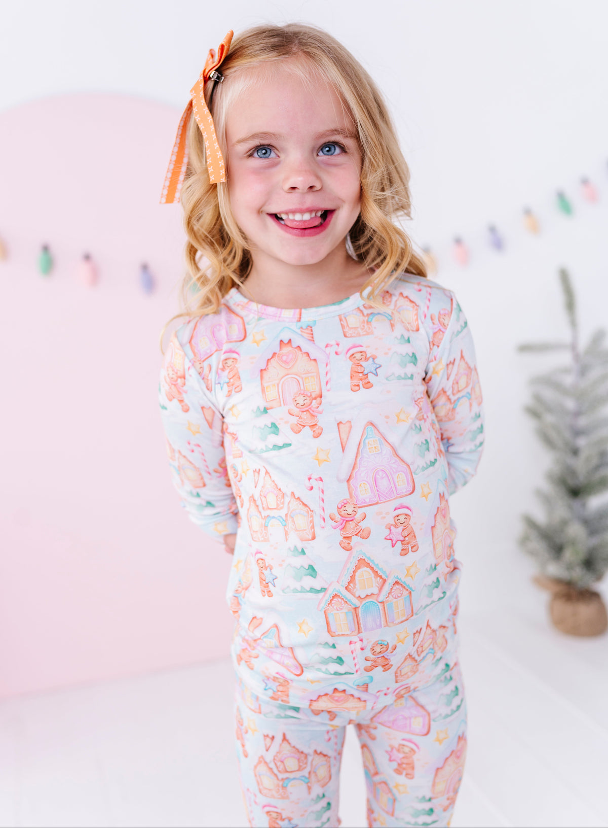 A young girl with blonde hair and a bow smiling while wearing a long-sleeve bamboo pajama set with a colorful gingerbread house pattern, set against a soft pink and white studio background.