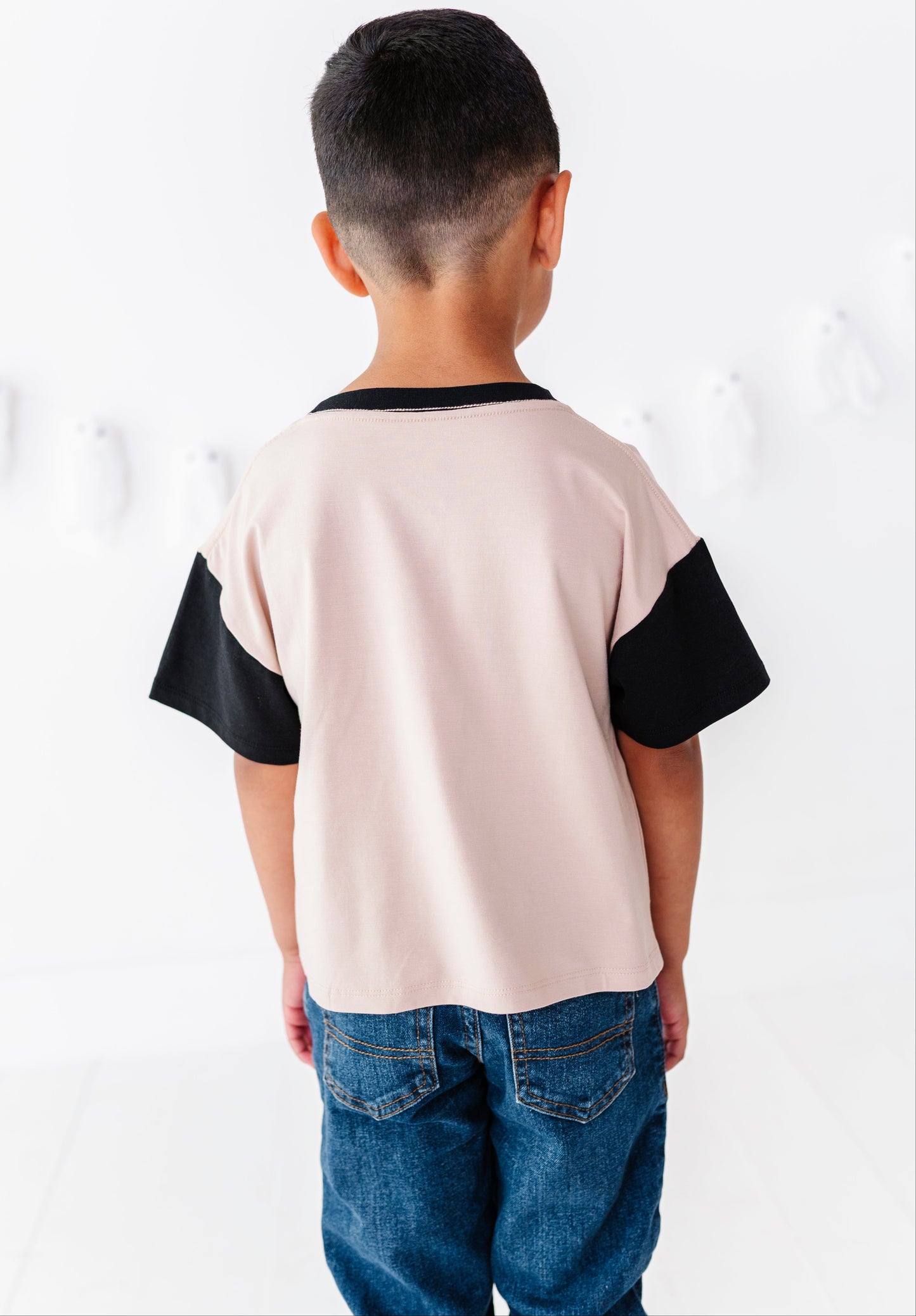 A child viewed from behind wearing a short-sleeve colorblock t-shirt with a light pink body and black sleeves, paired with blue jeans against a white studio background.