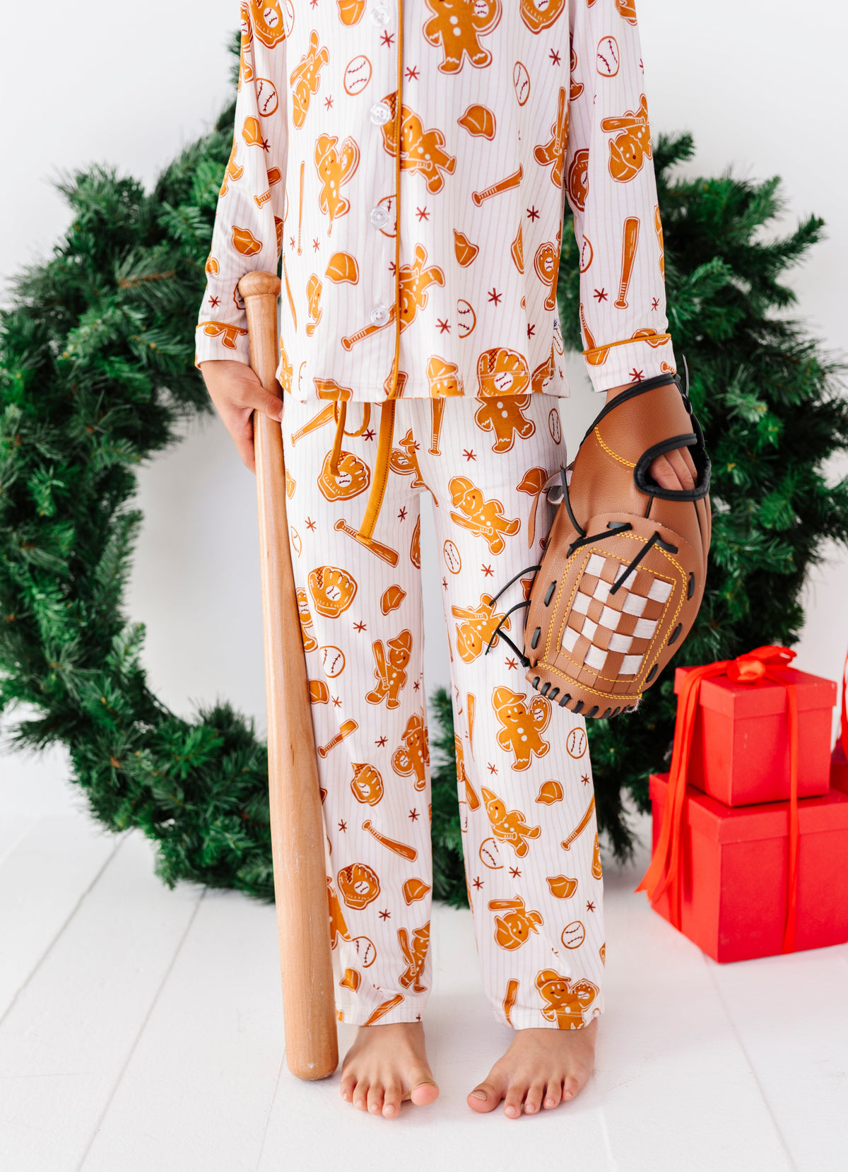 A child wearing white long-sleeve pajamas with an orange baseball-themed print, holding a wooden bat and wearing a baseball glove, standing in front of a green wreath with red wrapped gifts on a white floor.