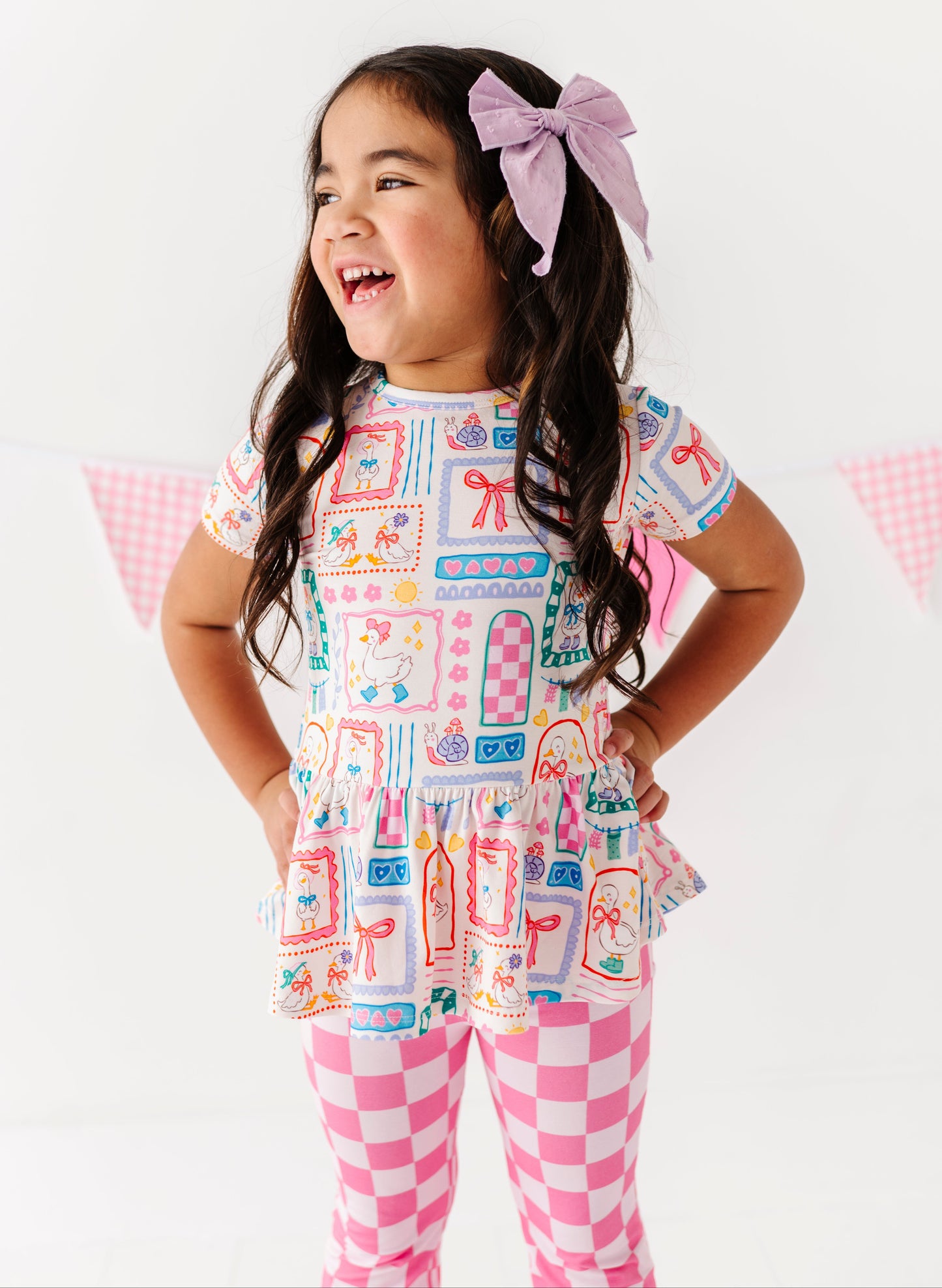 Young girl wearing a Gilly Goose Bamboo/Cotton Peplum tee with checkered flare pants on a white background