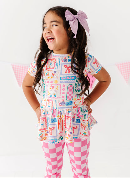 Young girl wearing a Gilly Goose Bamboo/Cotton Peplum tee with checkered flare pants on a white background