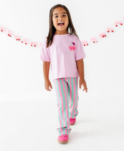A young girl walking forward in a studio wearing a light pink t-shirt with a cherry graphic and pastel pink and mint green striped pants, against a white background with a pink cherry garland.