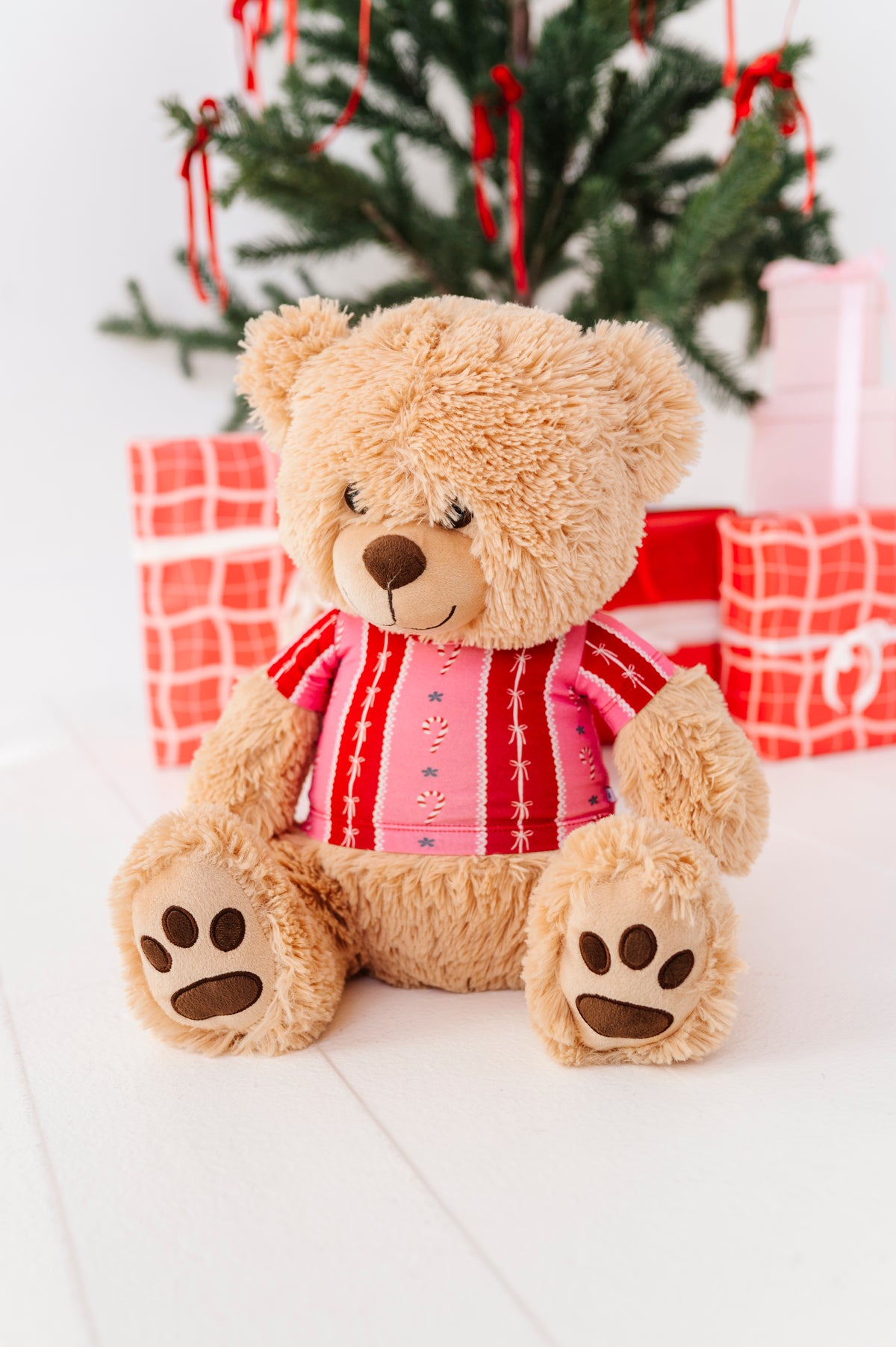 A plush teddy bear sits on a white surface in front of a decorated Christmas tree and wrapped gift boxes. The bear is tan and wears a pink and red striped bamboo shirt.