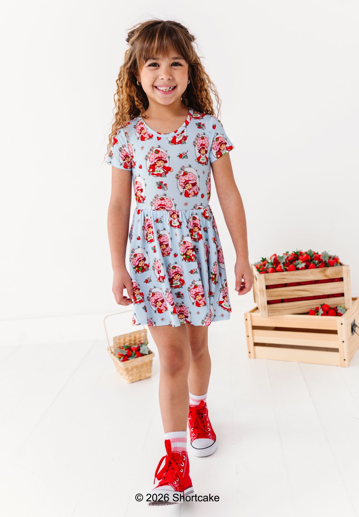 Young girl wearing a Strawberry Shortcake™ dress standing next to wooden crates filled with strawberries on a white background.