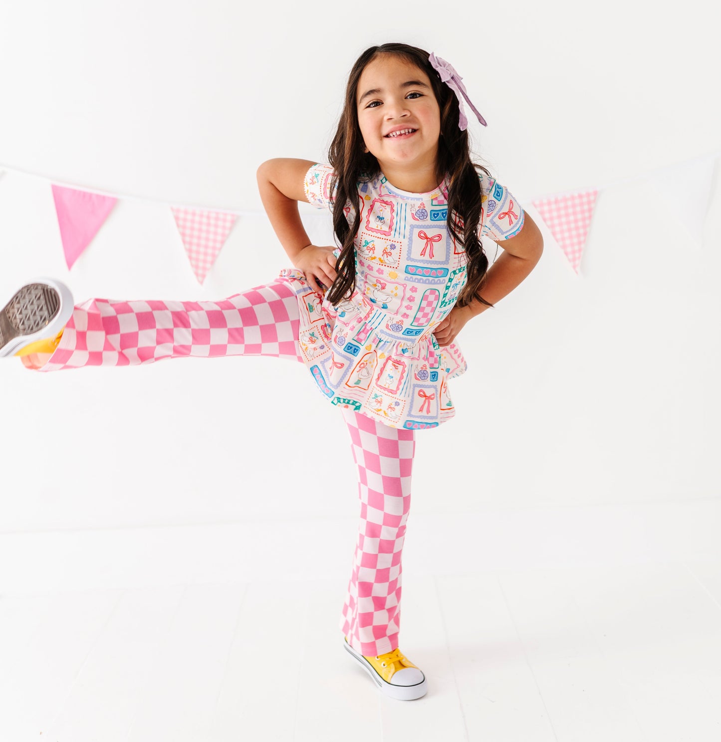 Young girl in a colorful Gilly Goose bamboo/cotton peplum top with pink checkered flare pants, standing against a white background.