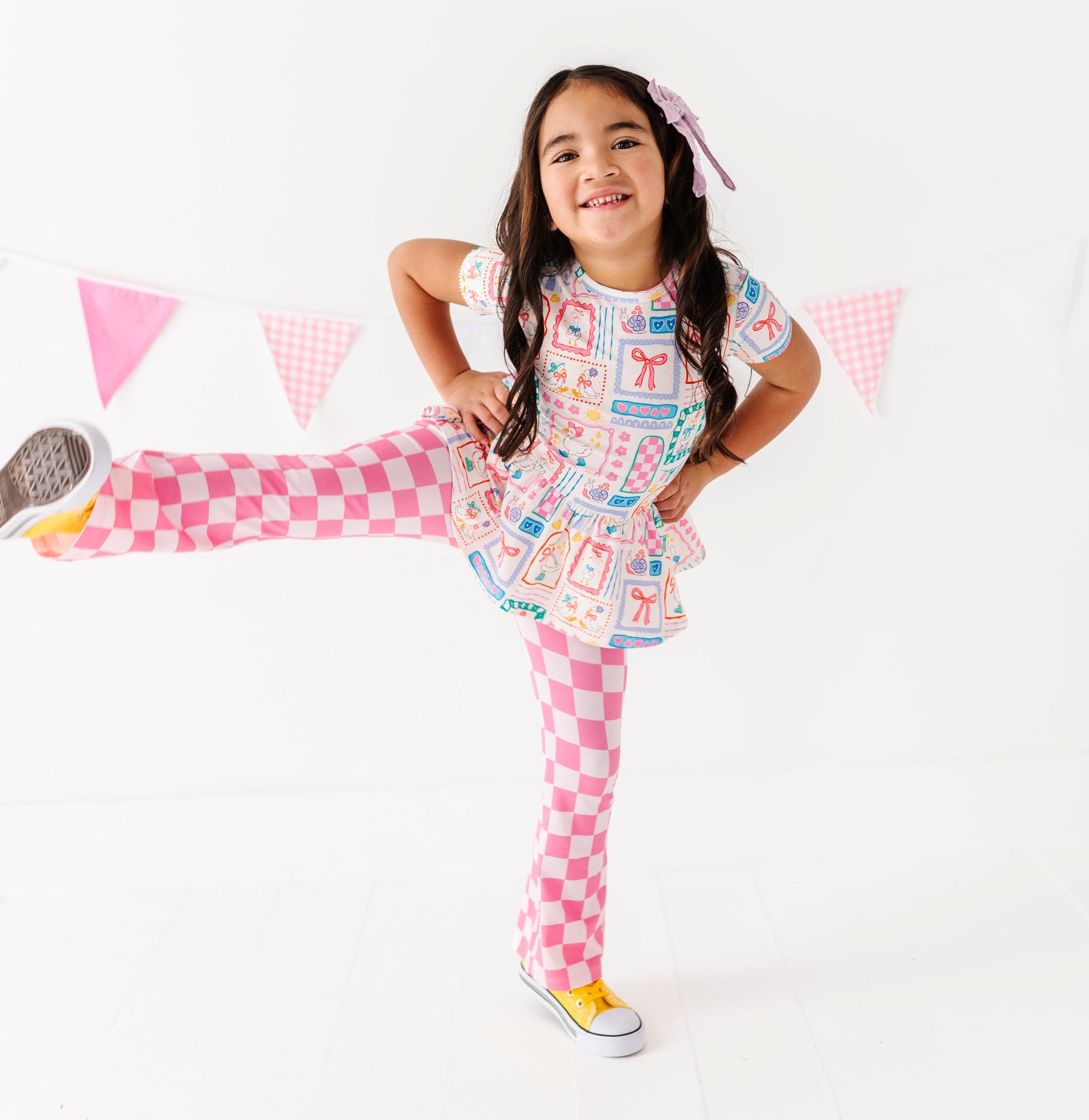 Young girl in a colorful Gilly Goose bamboo/cotton peplum top with pink checkered flare pants, standing against a white background.