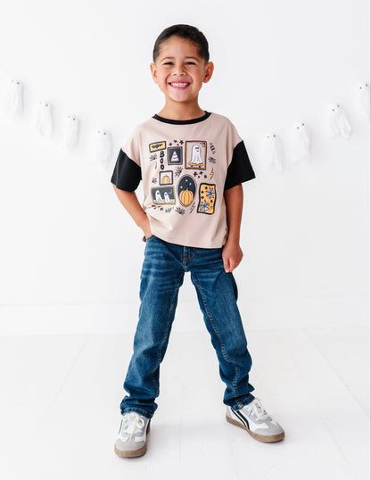 A smiling young boy stands in a bright white studio wearing a short-sleeve graphic t-shirt with beige and black panels, blue jeans, and white sneakers. Ghost decorations hang on the wall behind him.