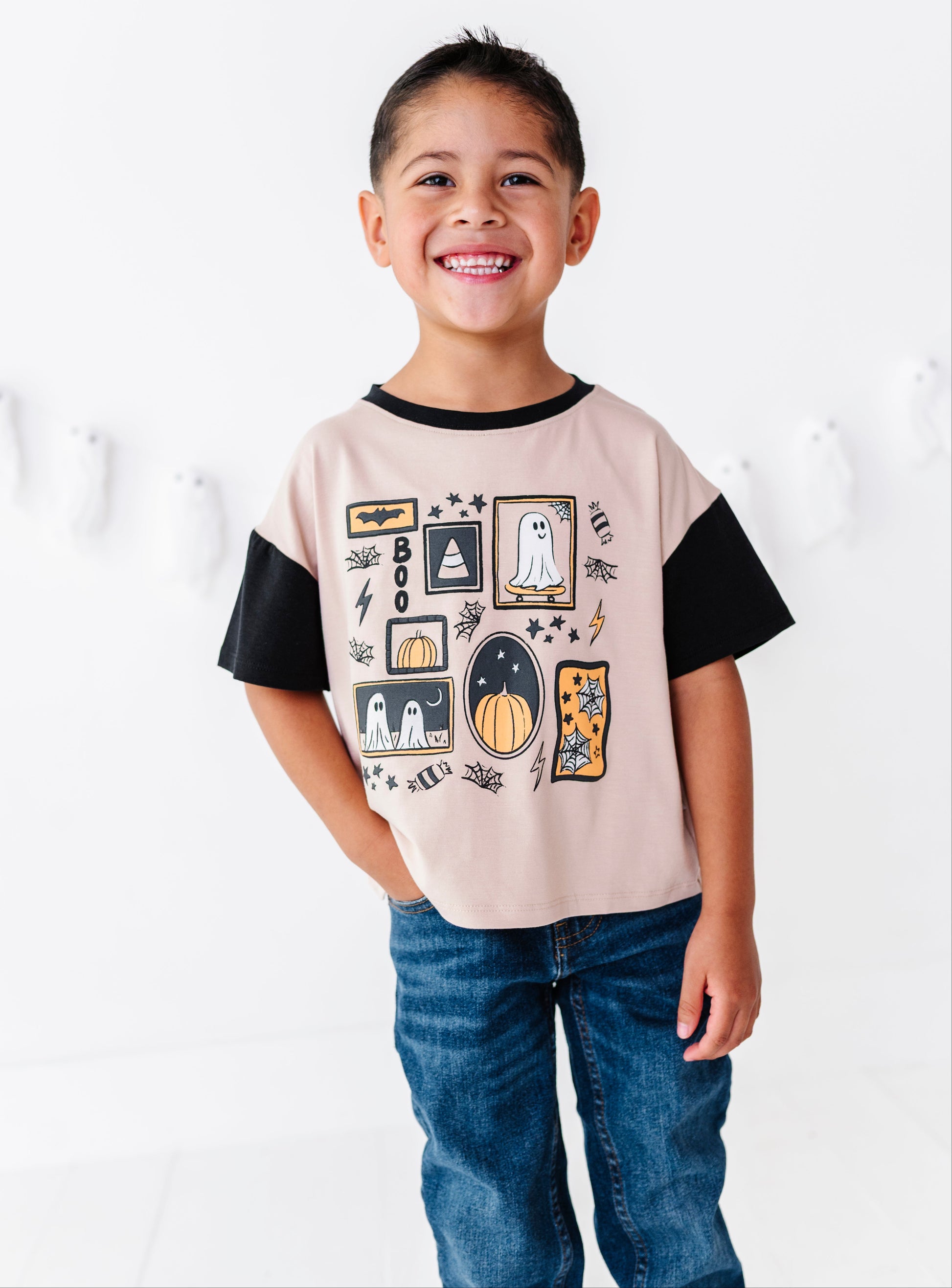 A smiling young boy wearing a short-sleeve t-shirt with a Halloween-themed graphic print, featuring black sleeves and a light pink body, paired with blue jeans against a white studio background.