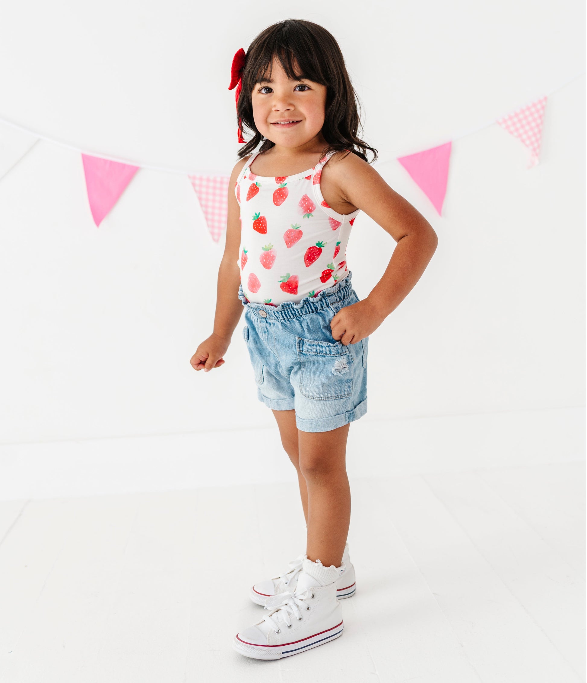 Young girl wearing a strawberry-patterned tank top and denim shorts, standing against a white background with pink triangle flags.