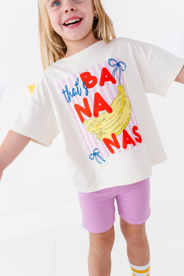 A young girl smiling and wearing a cream-colored graphic t-shirt with a colorful 'that's BANANAS' print featuring a banana illustration, paired with light purple shorts.
