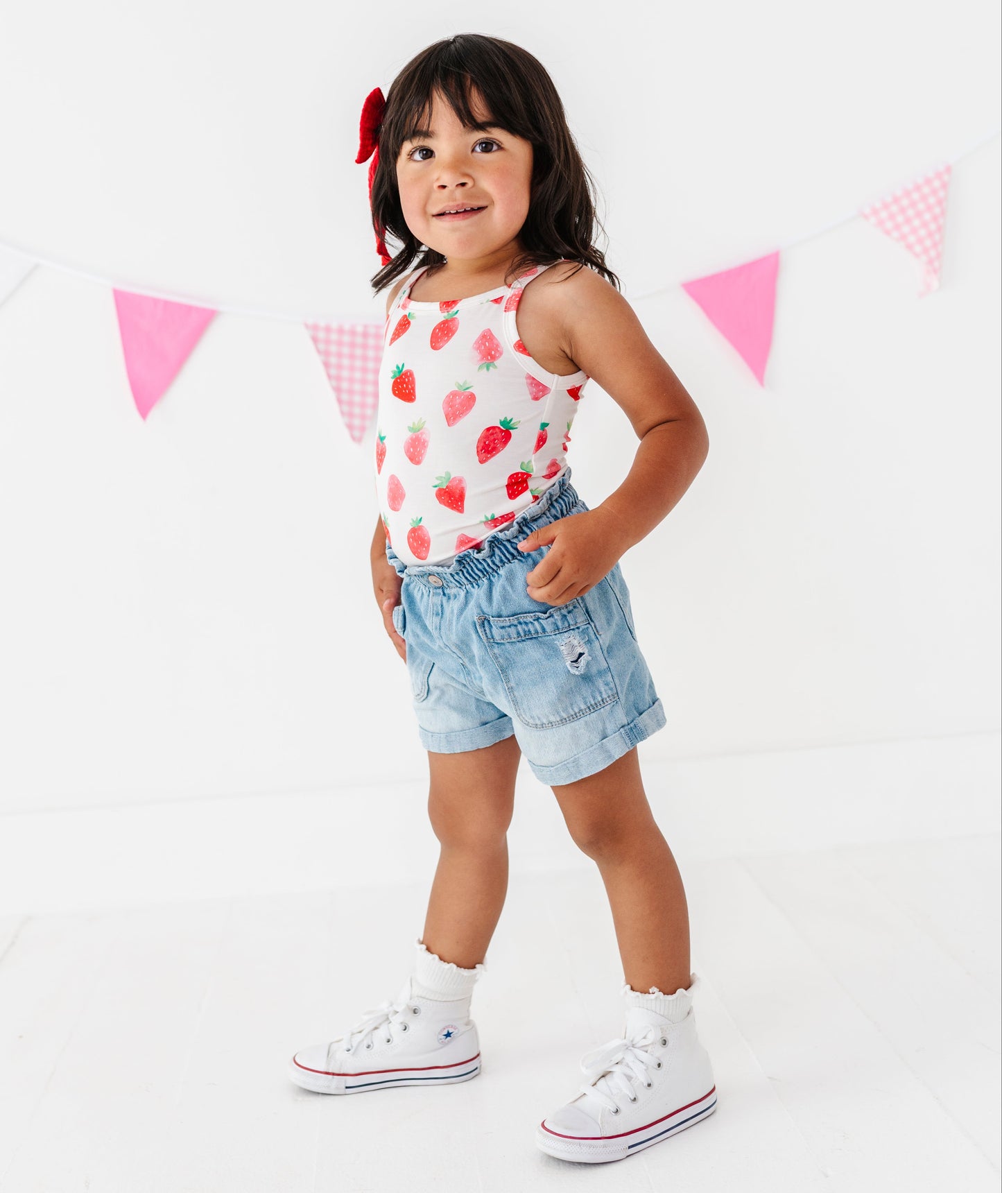 Young girl wearing a strawberry-patterned top and denim shorts standing in front of a pink triangle banner.