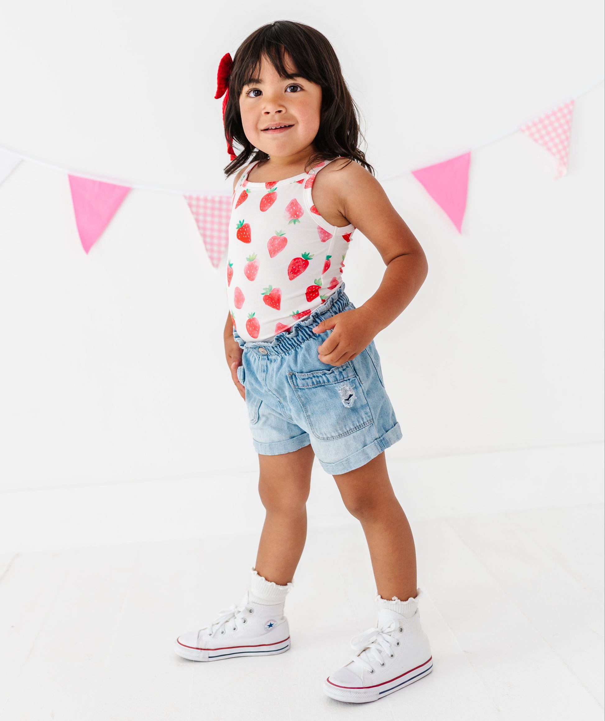 Young girl wearing a strawberry-patterned top and denim shorts standing in front of a pink triangle banner.