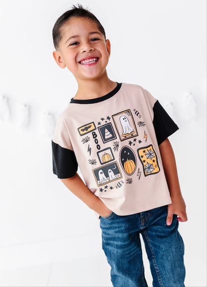A smiling young boy wearing a short-sleeve t-shirt with black sleeves and a beige body, decorated with a Halloween graphic print, paired with blue jeans against a white background.