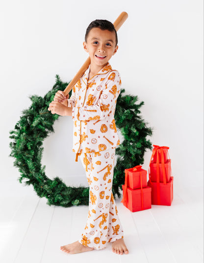 A young boy stands barefoot in holiday-themed pajamas, holding a wooden baseball bat over his shoulder. He is posed in front of a green Christmas wreath with red wrapped presents on a clean white background.