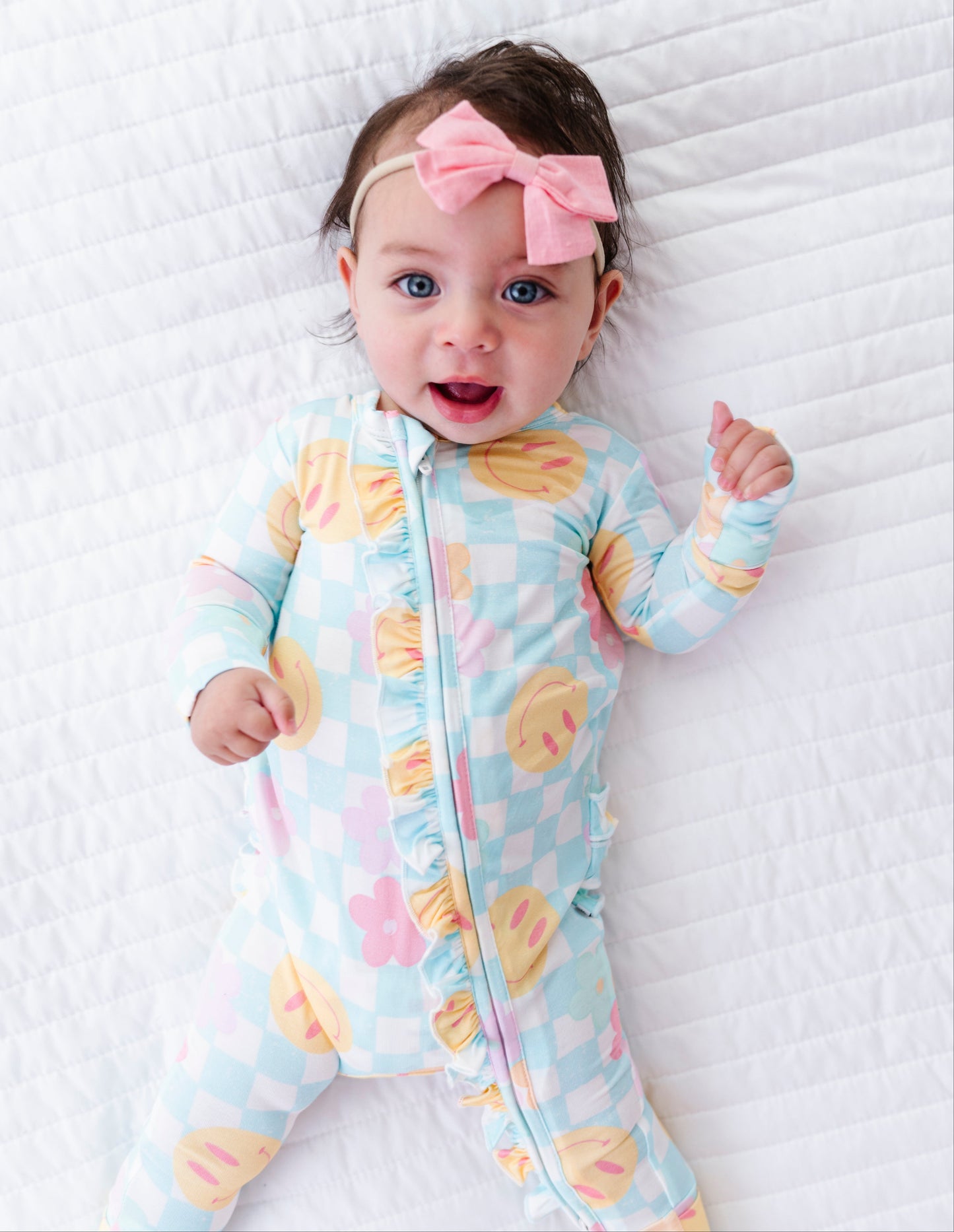 A baby lying on a white quilted blanket, wearing a light blue and white checkered footed sleeper with pastel macaron and flower prints, along with a pink bow headband.
