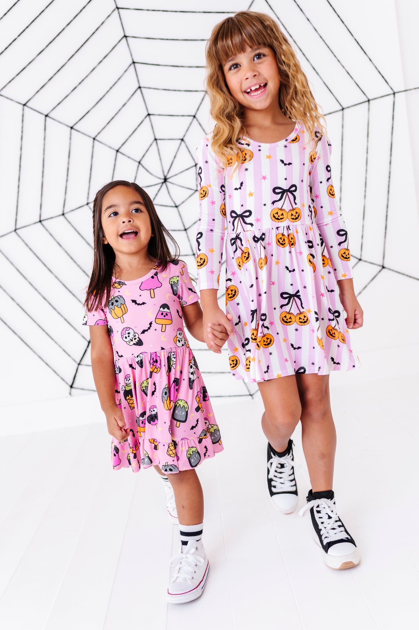 Two young girls wearing matching pink and white long-sleeve dresses with Halloween prints, standing in front of a black spiderweb backdrop on a white studio background.