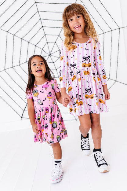 Two young girls wearing matching pink and white long-sleeve dresses with Halloween prints, standing in front of a black spiderweb backdrop on a white studio background.
