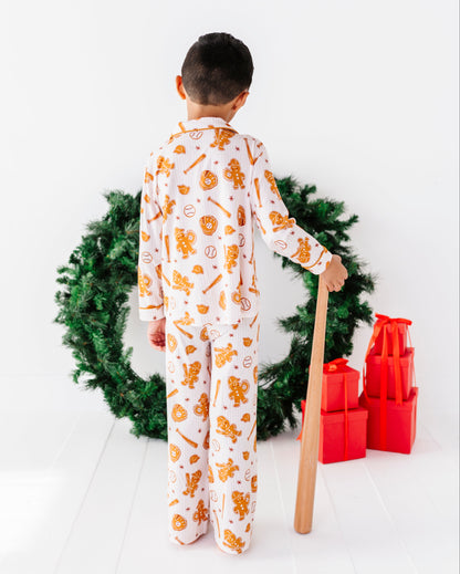A child stands with their back to the camera wearing a white long-sleeve pajama set with an orange gingerbread-themed print. They are holding a wooden baseball bat next to a green Christmas wreath and red wrapped gifts on a white wooden floor.