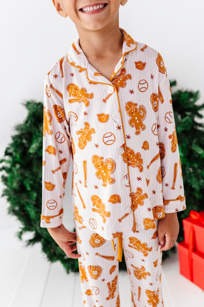 A child wearing a matching long-sleeve pajama set with an orange baseball-themed print, including baseballs, bats, and a gingerbread person, shown against a festive holiday background with a Christmas tree and red gift boxes.