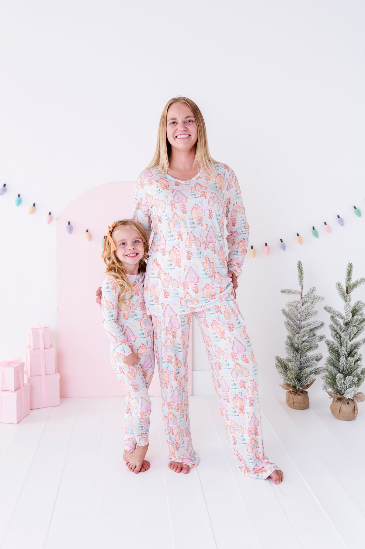 A woman and a young girl wearing matching pastel pajamas with a whimsical unicorn pattern, standing in a bright, white room decorated with a pink arch, string lights, and small potted trees.