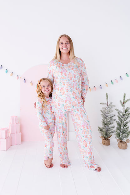 A woman and a young girl wearing matching pastel pajamas with a whimsical unicorn pattern, standing in a bright, white room decorated with a pink arch, string lights, and small potted trees.
