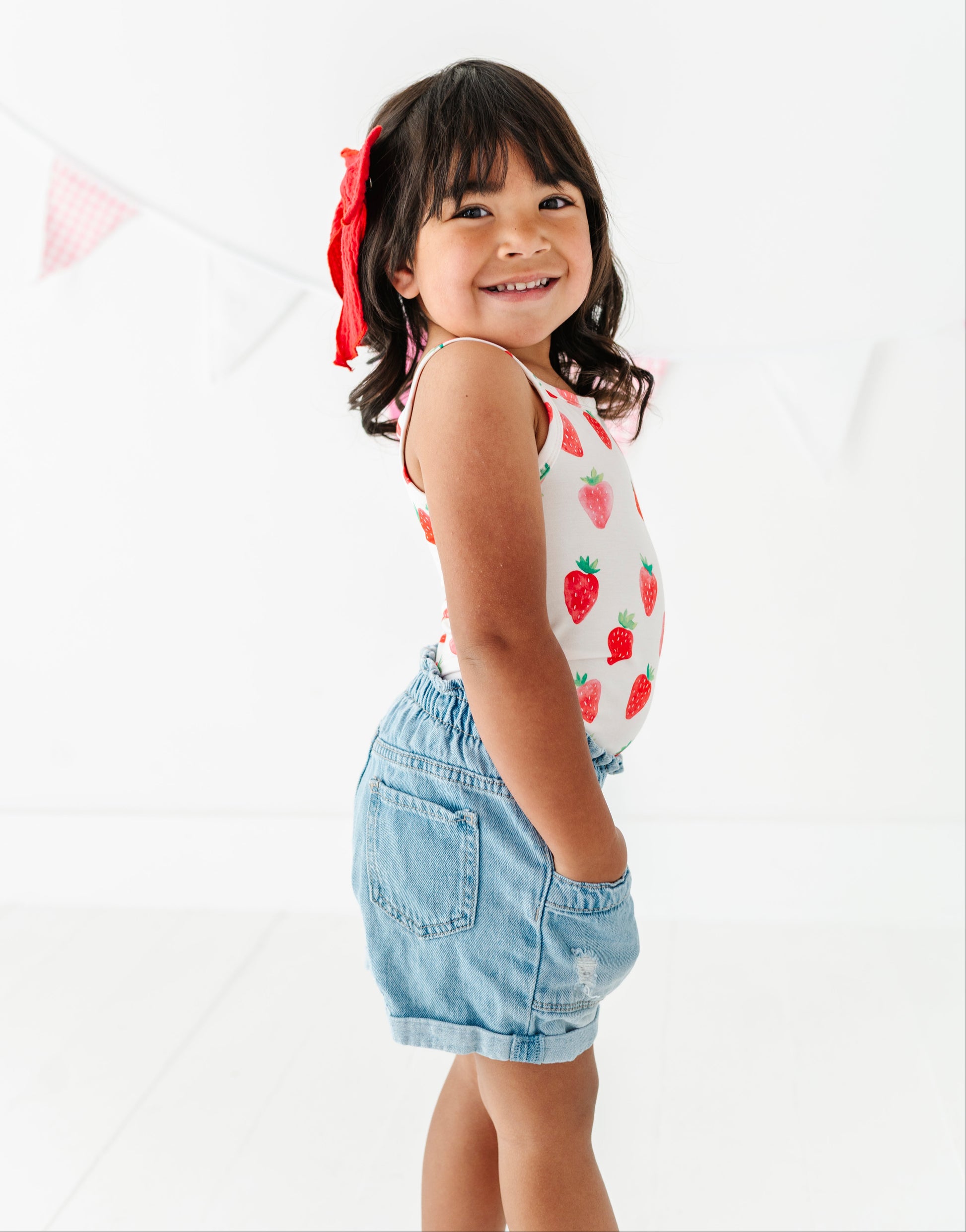 Young girl wearing a tank top with strawberry pattern and denim shorts on a white background