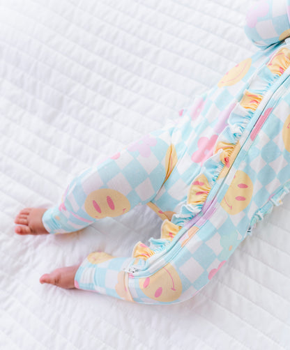 A close-up of a baby's legs wearing a pastel checkered footed sleeper with ruffle trim, lying on a white quilted blanket.