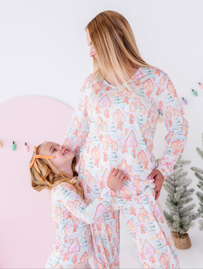 A woman and a young girl wearing matching long-sleeve bamboo pajamas with a pastel gingerbread house pattern, standing in a bright room with a pink wall and a small tree.