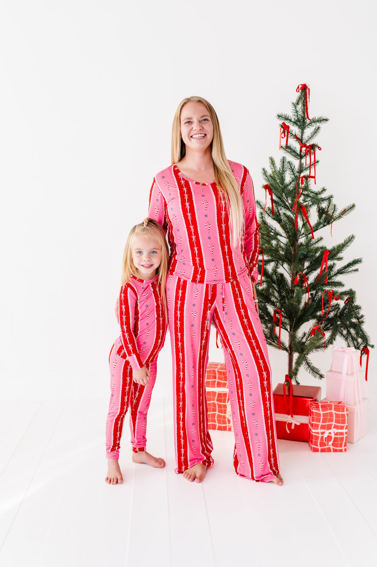 A woman and a young girl wearing matching pink and red striped bamboo Christmas pajamas, standing barefoot in front of a decorated Christmas tree with wrapped gifts on a white studio floor.
