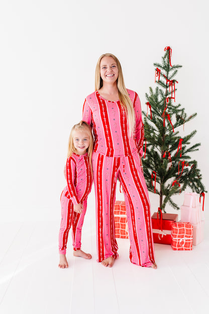 A woman and a young girl wearing matching pink and red striped bamboo Christmas pajamas, standing barefoot in front of a decorated Christmas tree with wrapped gifts on a white studio floor.