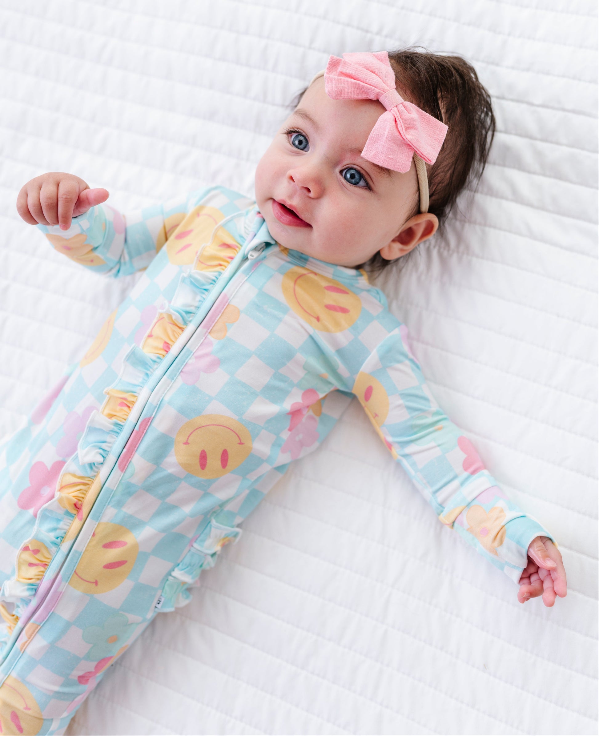 A baby lying on a white quilted blanket, wearing a light blue and white checkered footed sleeper with yellow smiley face prints and a pink bow headband.