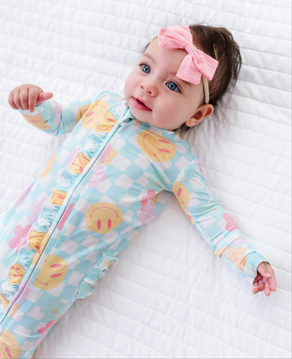 A baby lying on a white quilted blanket, wearing a light blue and white checkered footed sleeper with yellow smiley face prints and a pink bow headband.