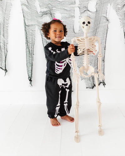 Child in skeleton-themed outfit holding a small skeleton model against a Halloween-themed backdrop.