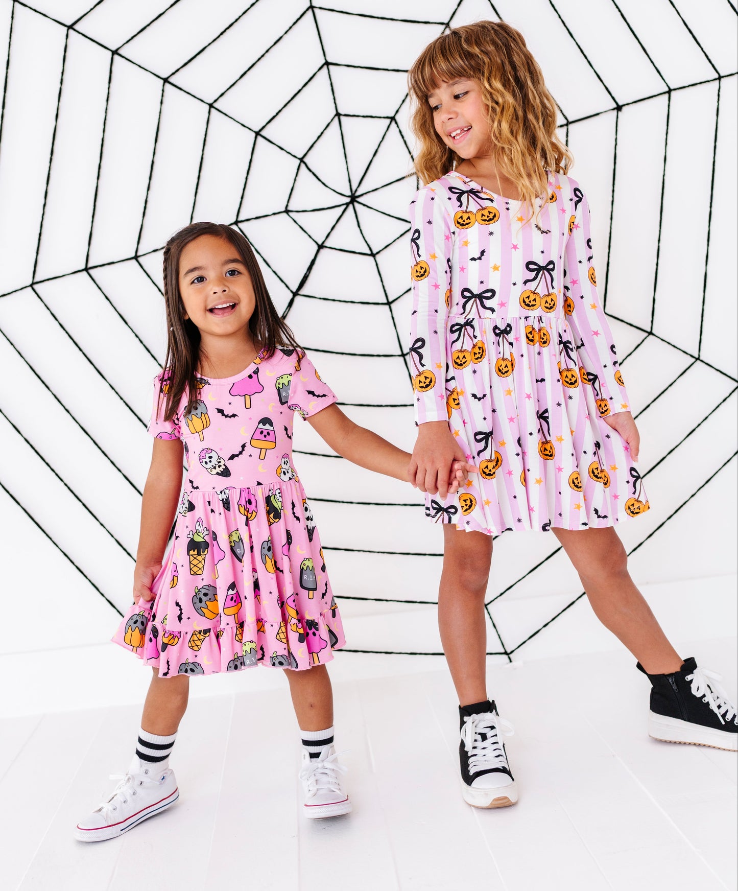 Two young girls wearing matching pink patterned dresses stand holding hands in front of a large black spiderweb graphic on a white background.