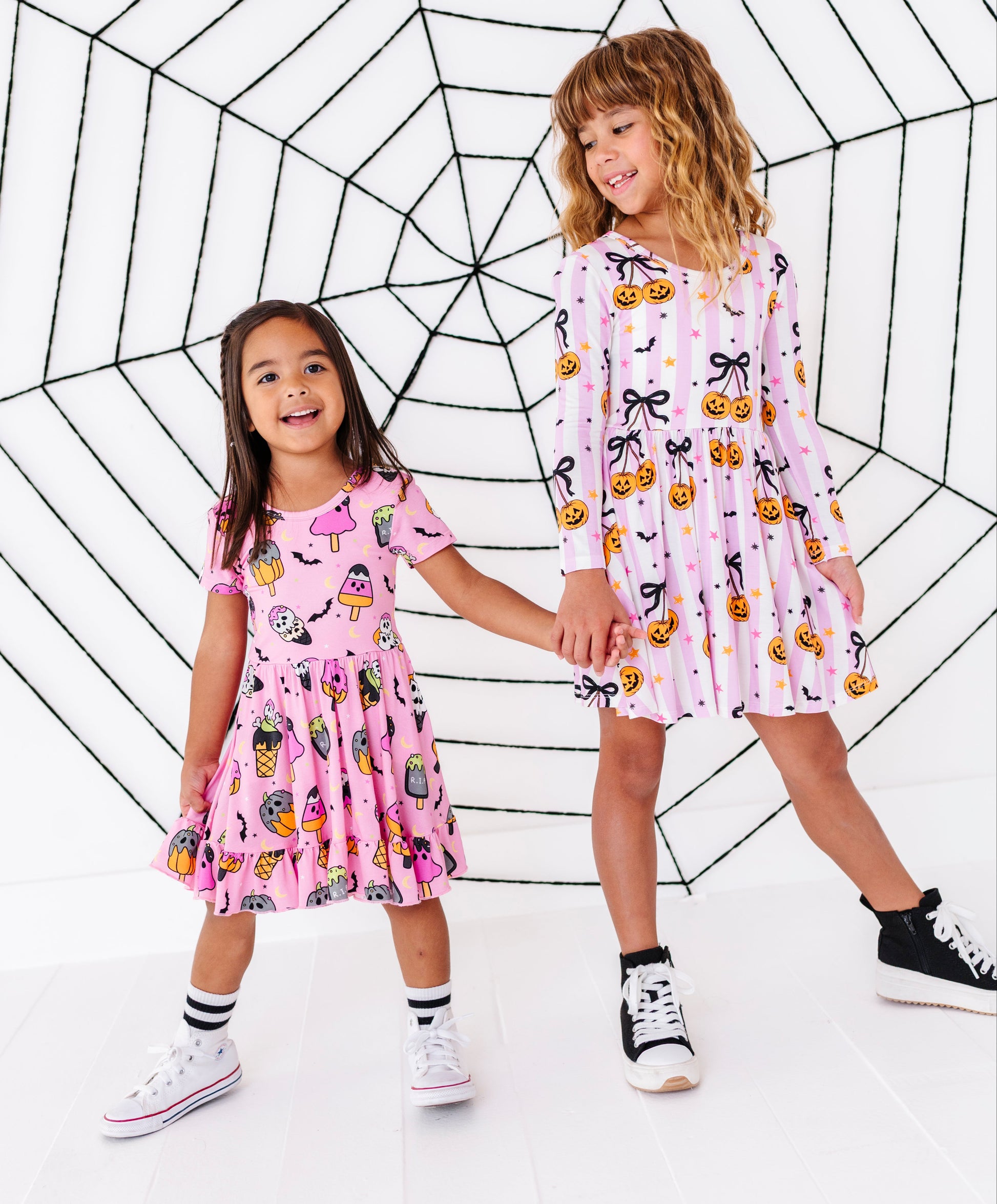 Two young girls wearing matching pink patterned dresses stand holding hands in front of a large black spiderweb graphic on a white background.