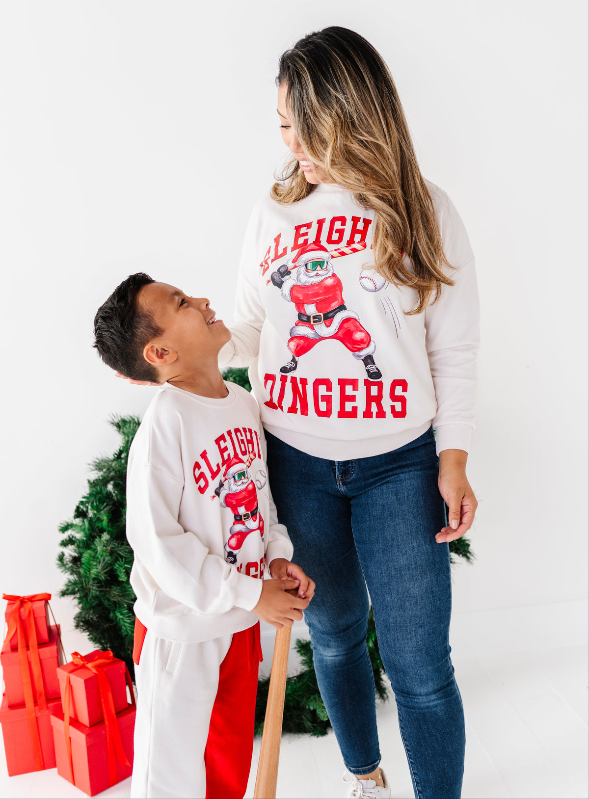 A woman and a young boy wearing matching white Christmas sweaters with a red and black hockey player graphic and the words ' sleighing jingers' pose in front of a white studio background with a small green tree and red gift boxes.