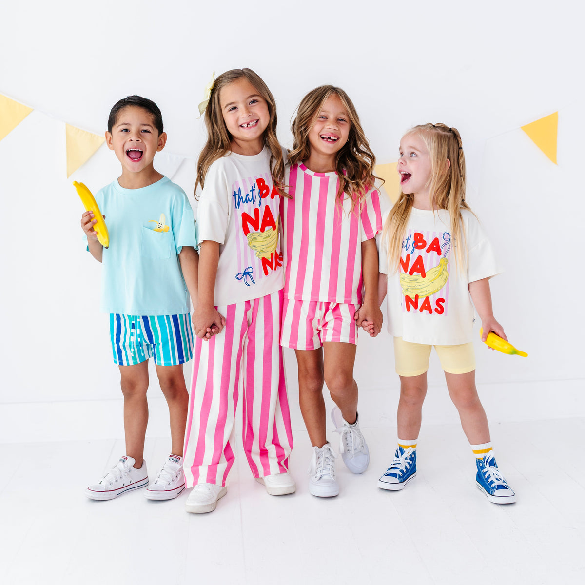 Four children standing together in a bright studio setting, each wearing a colorful graphic T-shirt and matching shorts or pants. The outfits feature playful prints and patterns including blue/yellow, pink/white stripes, and cream/yellow graphics.